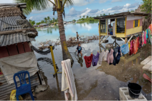 Fotografia mostranco região pobre e alagada. Duas crianças brincam na água poluída. Há roupas no varal e casas pequenas sustentadas por madeiras.